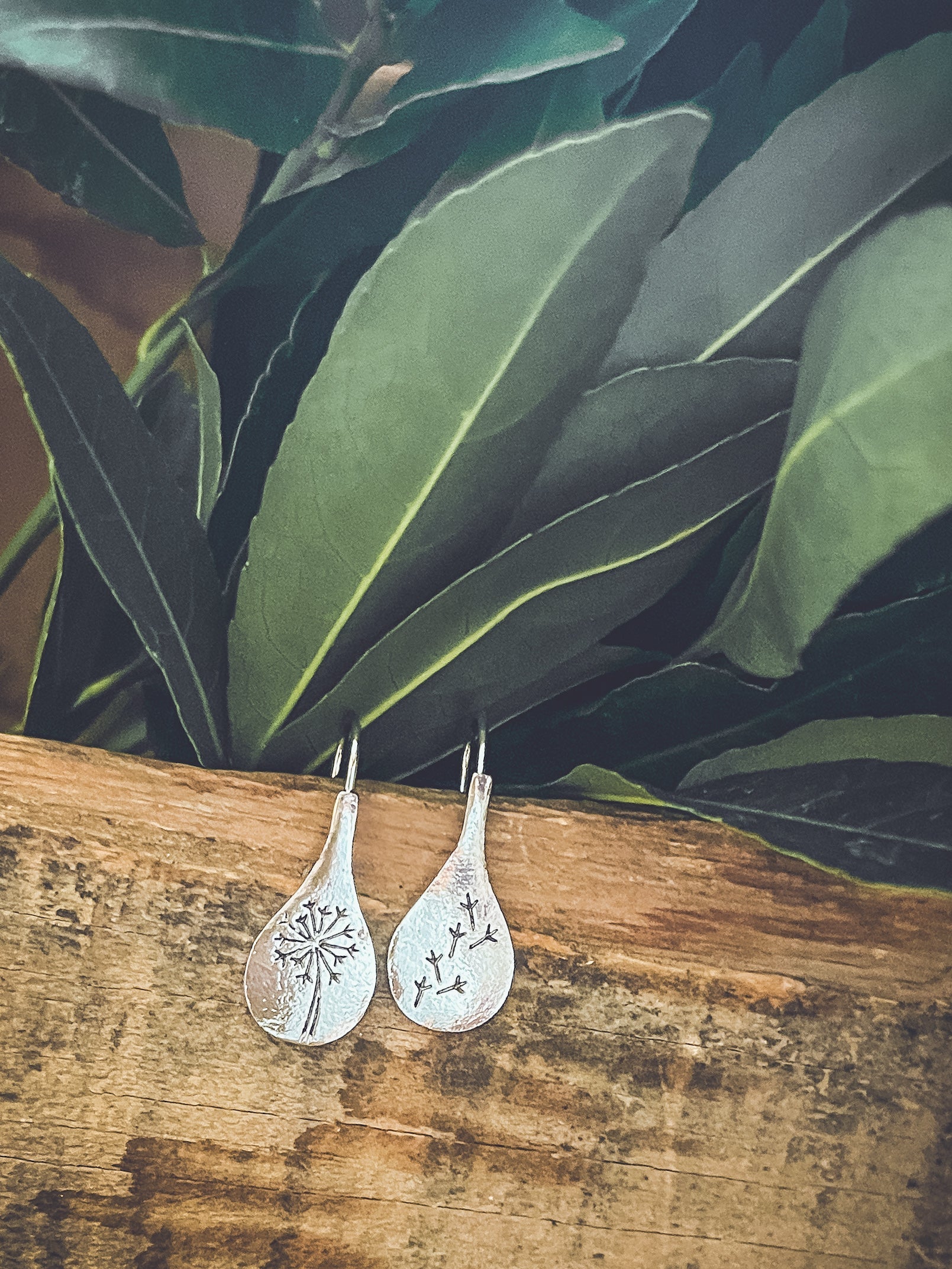 Silver teardrop earrings with engraved designs on a wooden surface with green leaves in the background