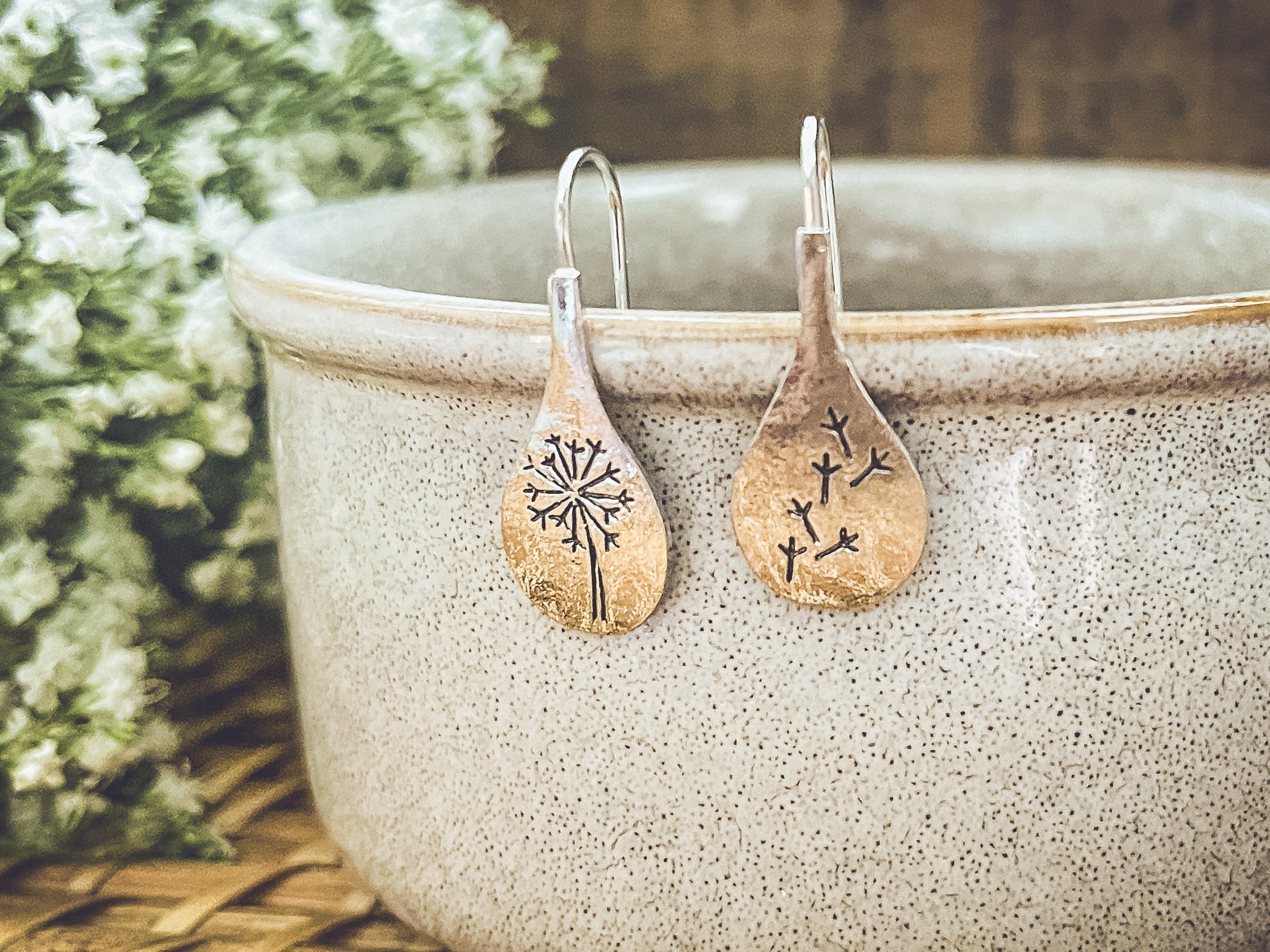 Teardrop-shaped earrings with tree design on a ceramic bowl with flowers in the background